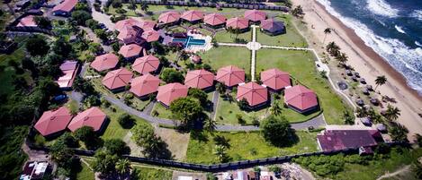 Vista aérea de un resort con varias edificaciones de techo rojo, una piscina central y acceso a la playa.