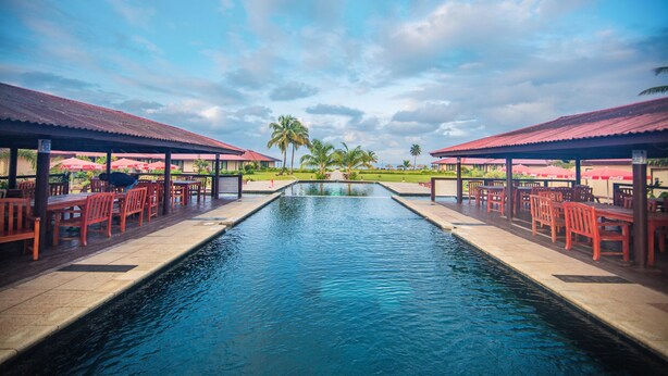 A swimming pool with red chairs and umbrellas, surrounded by a covered area with palm trees in the background. - Rlj Kendeja Resort and Villas (Monrovia)