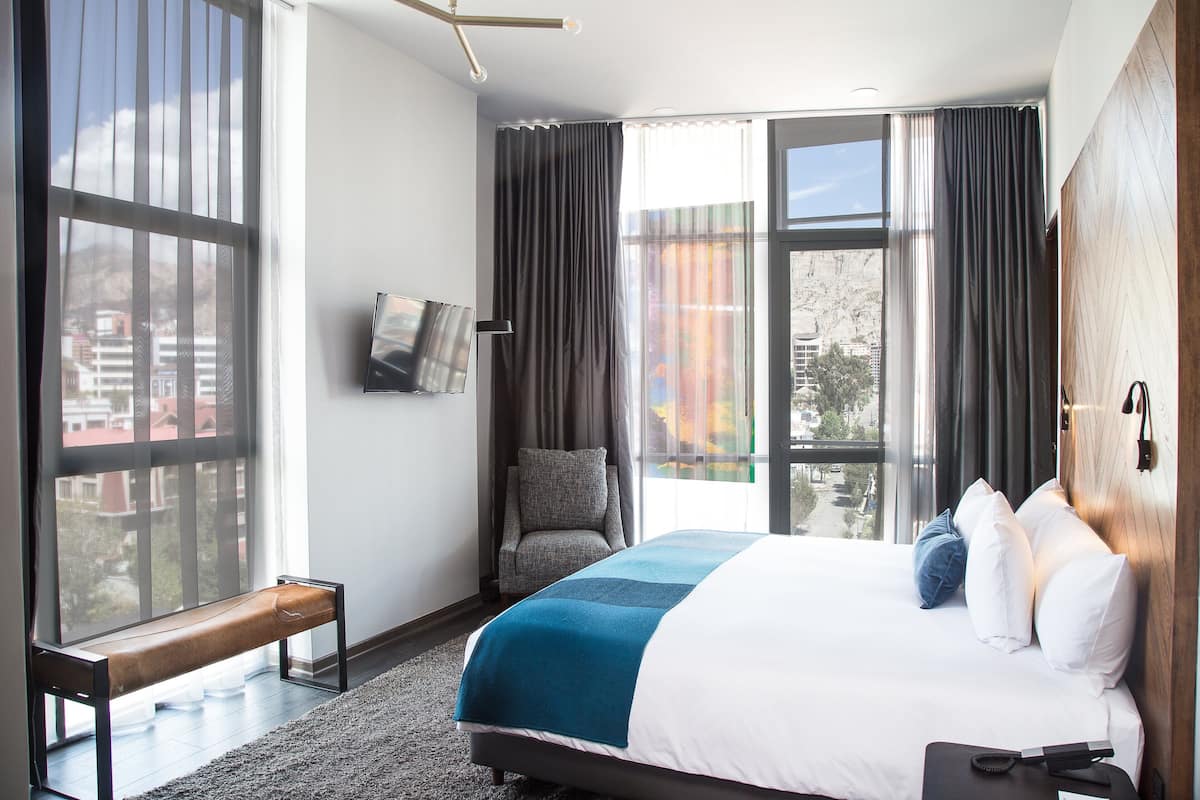 A modern hotel room with a large bed, a bench, a chair, and a view of the city through large windows.