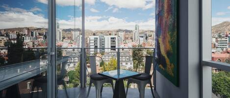 Un balcón con baranda de vidrio, una mesa con dos sillas y un cuadro colorido en la pared.