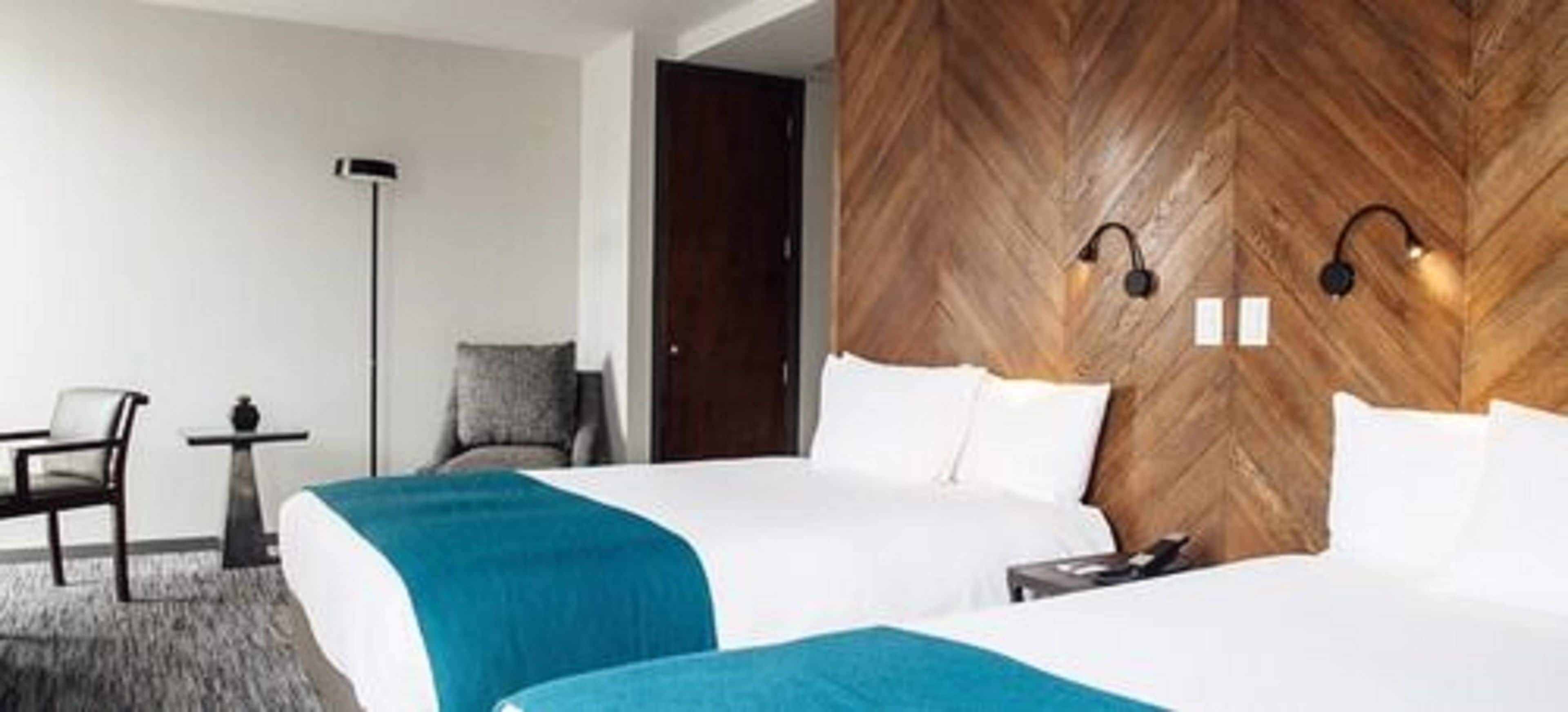 A hotel room with two beds, a desk, a chair, and a wooden accent wall with a light fixture.