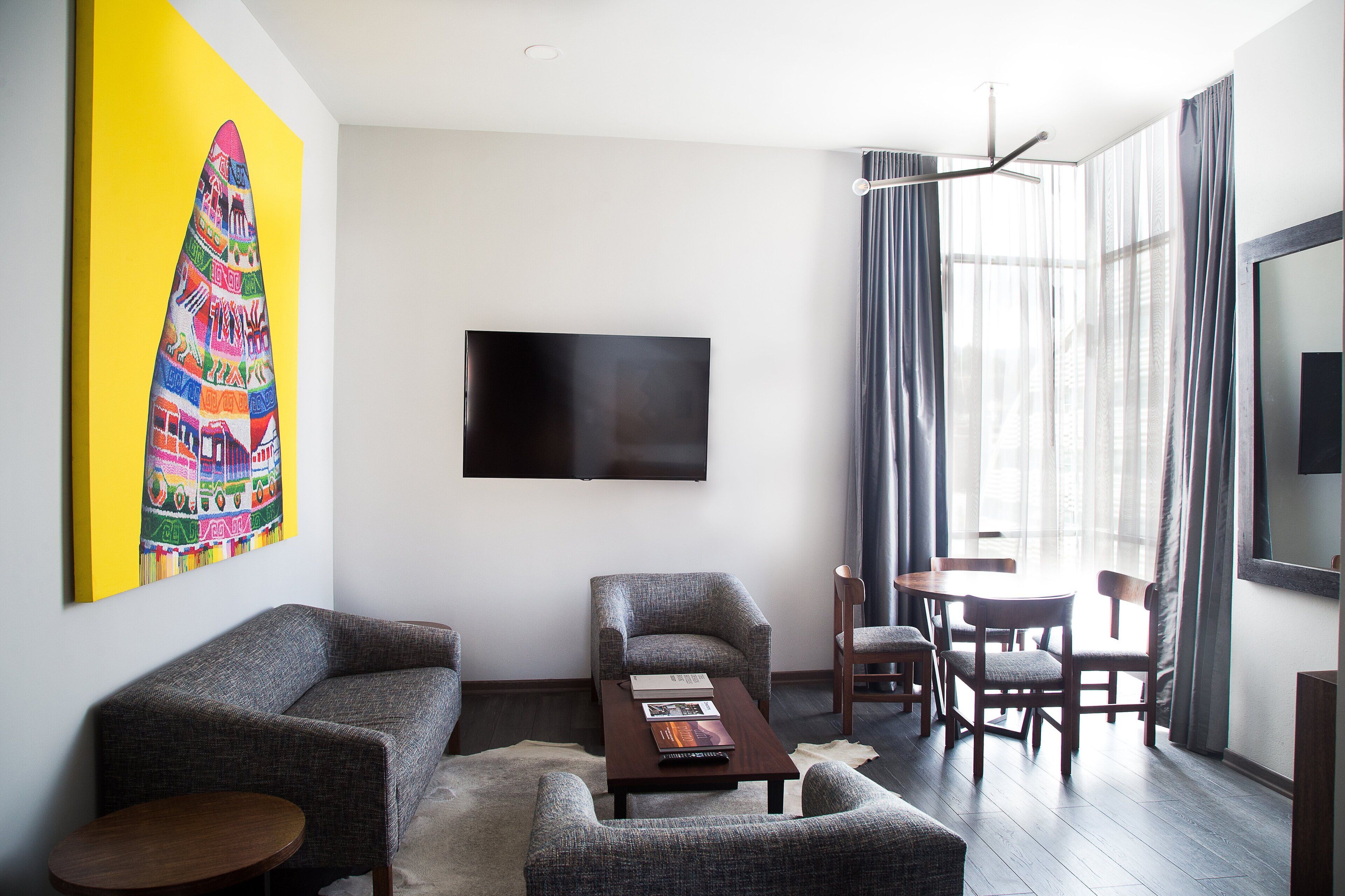 A modern living room with a sofa, armchairs, a coffee table, and a dining area with a table and chairs. A flat-screen TV is mounted on the wall, and a colorful artwork is displayed above the sofa.