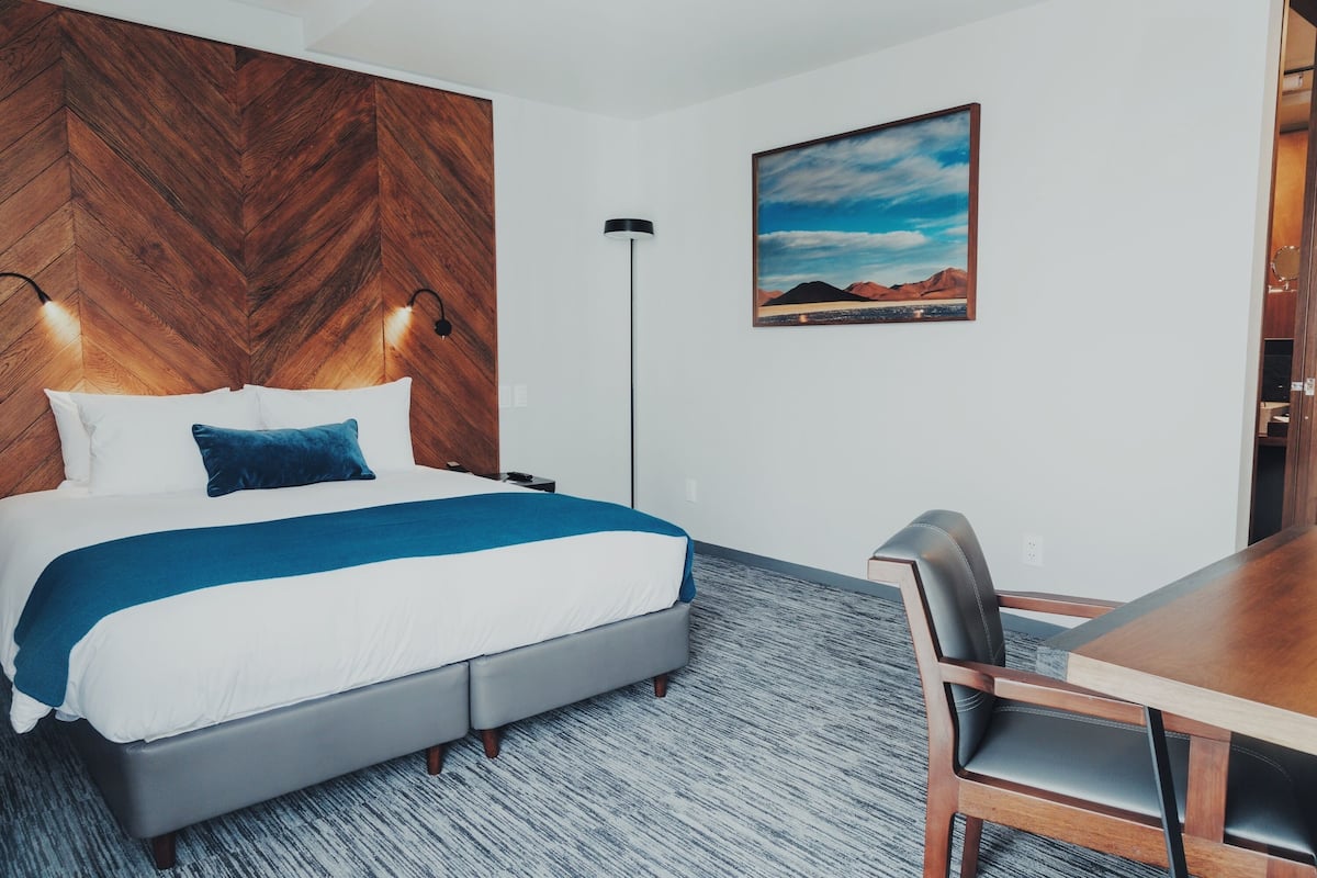 A hotel room with a bed, a desk, a chair, a lamp, and a painting on the wall.