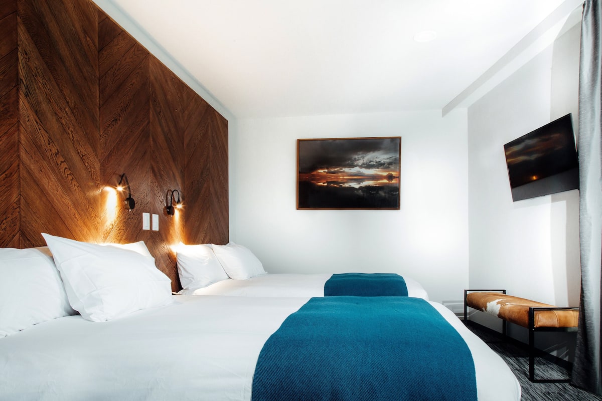 A modern hotel room with a large bed, a wooden headboard, a mounted painting, and a flat-screen TV.