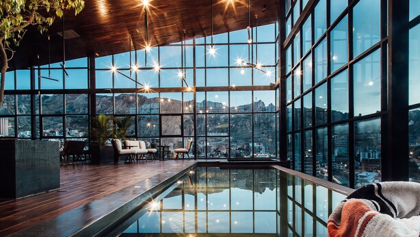 Piscina interior moderna con barrera de vidrio, suelo de madera y vistas a un paisaje urbano y montañas.