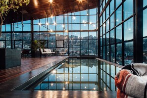 Une piscine intérieure moderne, dotée d’une paroi en verre, d’un plancher en bois et offrant une vue sur le paysage urbain et les montagnes.
