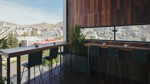 Zona de bar moderna con mostrador de madera y sillas negras, con vistas a un paisaje urbano y montañas al fondo.