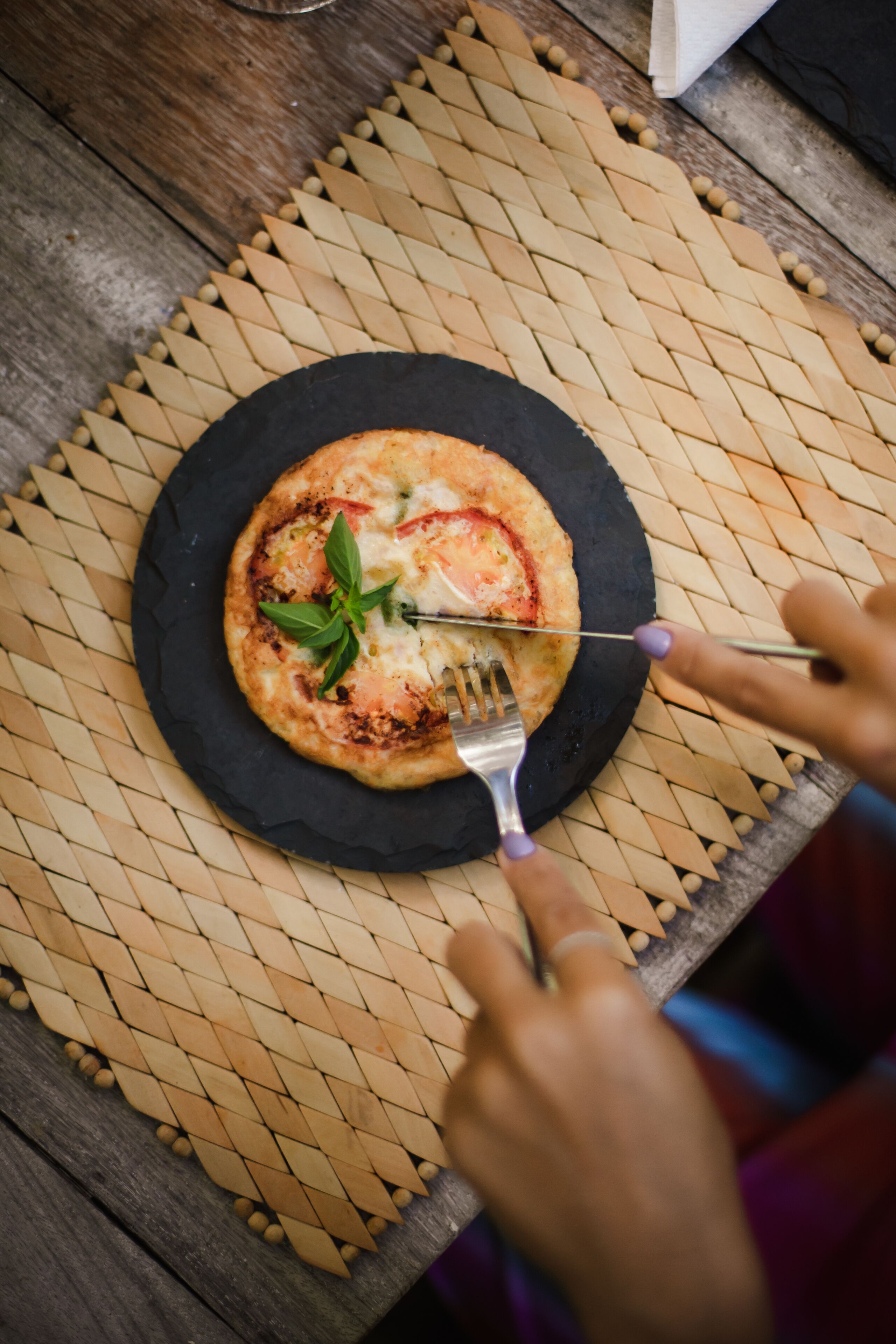 En rund pizza på en svart tallrik, med en gaffel bredvid, placerad på en vävd matta.