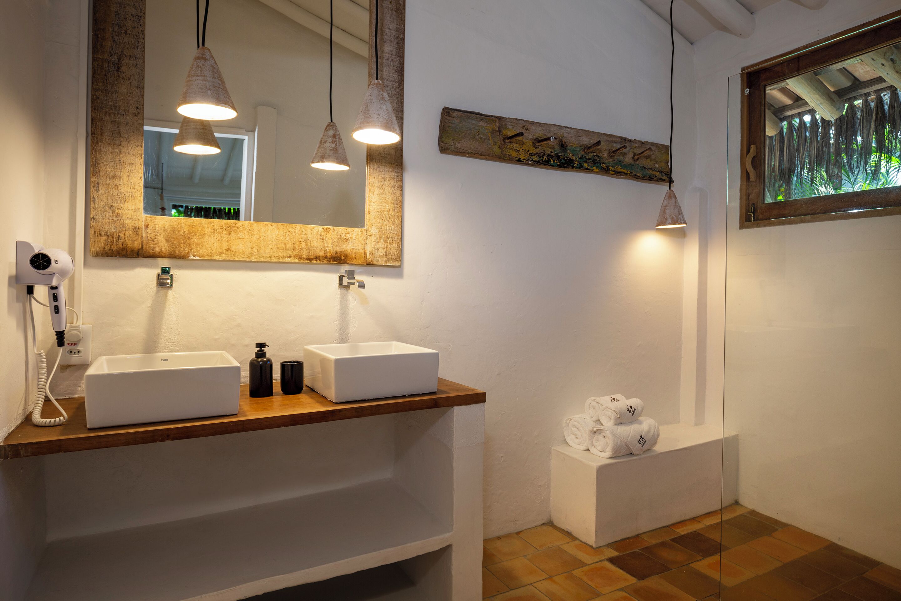A modern bathroom with two sinks, a large mirror, pendant lights, a hairdryer, and a window with a view of greenery.