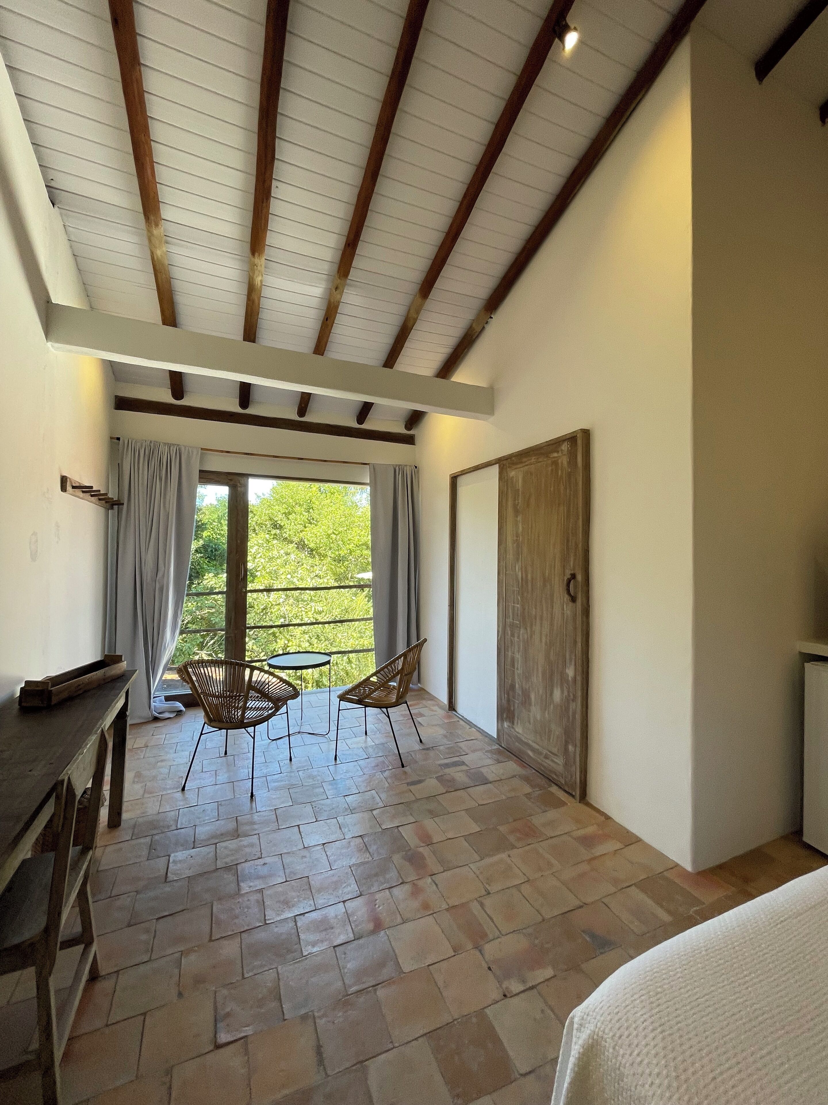 Quarto com teto alto, vigas de madeira expostas, grande porta de vidro deslizante para uma varanda, pequena mesa redonda com duas cadeiras de vime e um banco de madeira.