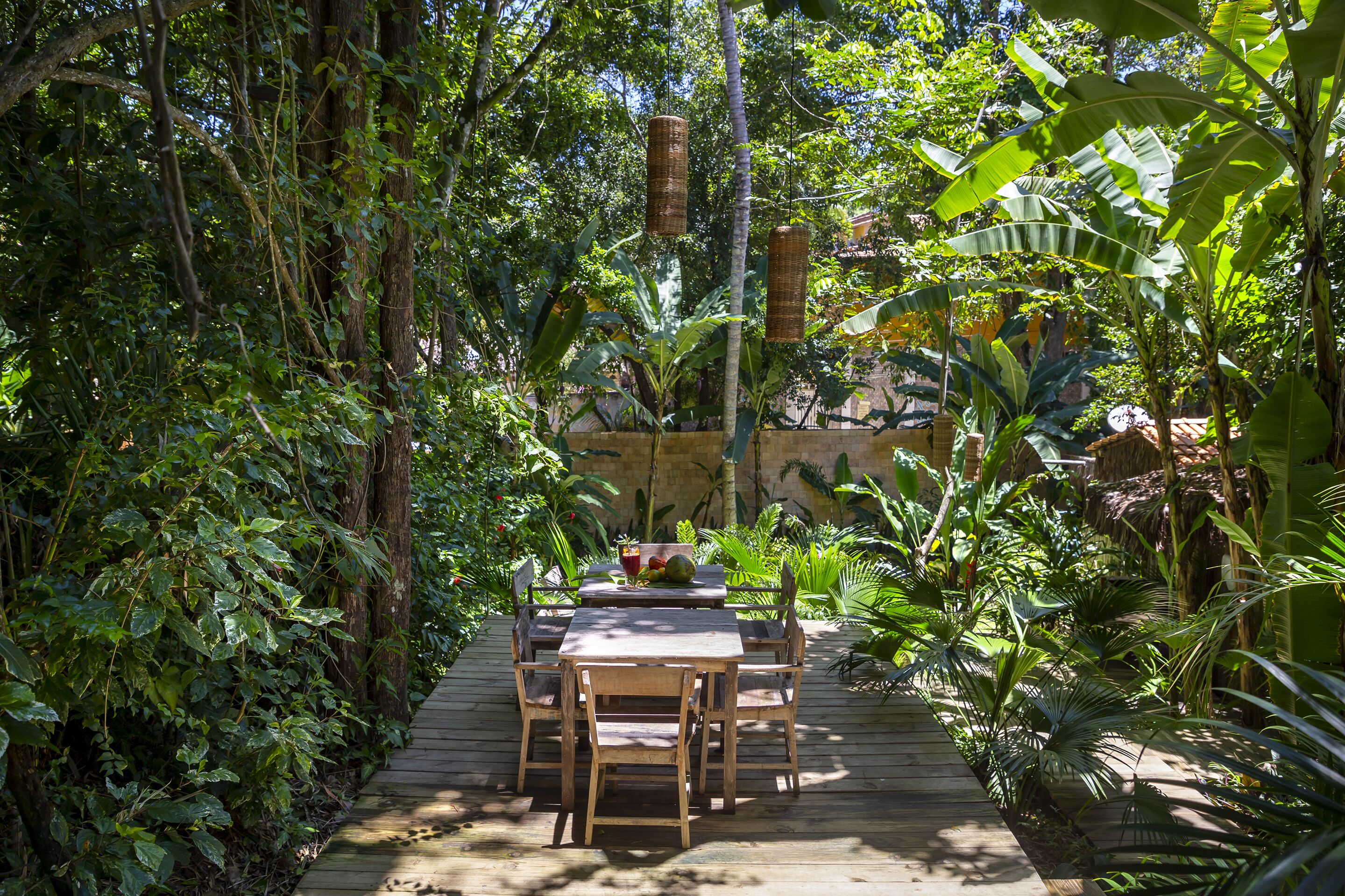 A wooden deck with a table and chairs surrounded by lush greenery and a stone wall.