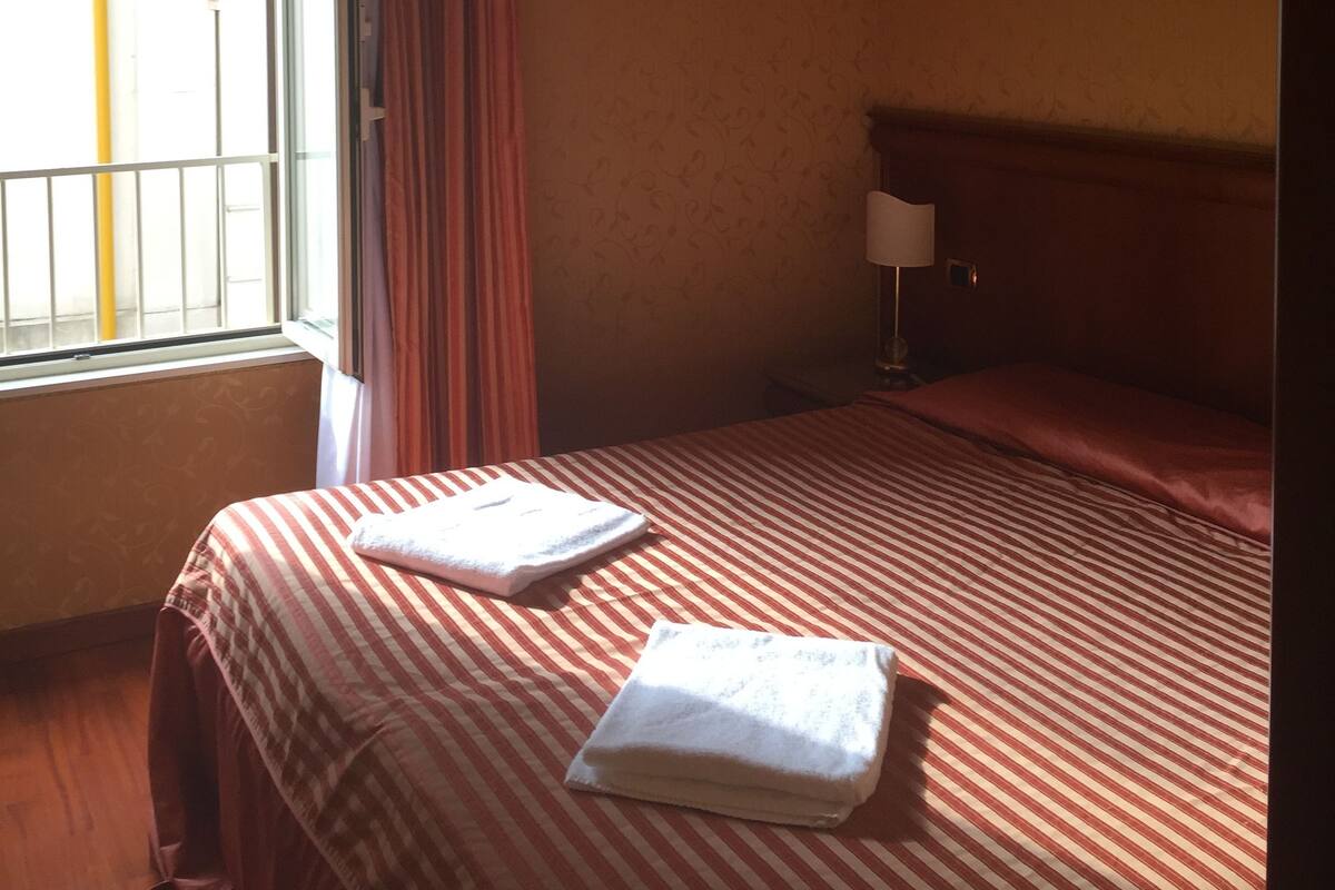 A hotel room with a bed, a nightstand, a window with curtains, and a view of the outside.