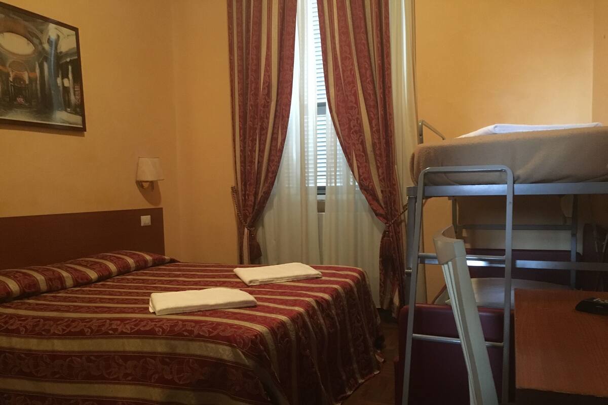 A hotel room with two beds, a bunk bed, a painting on the wall, and a window with curtains.