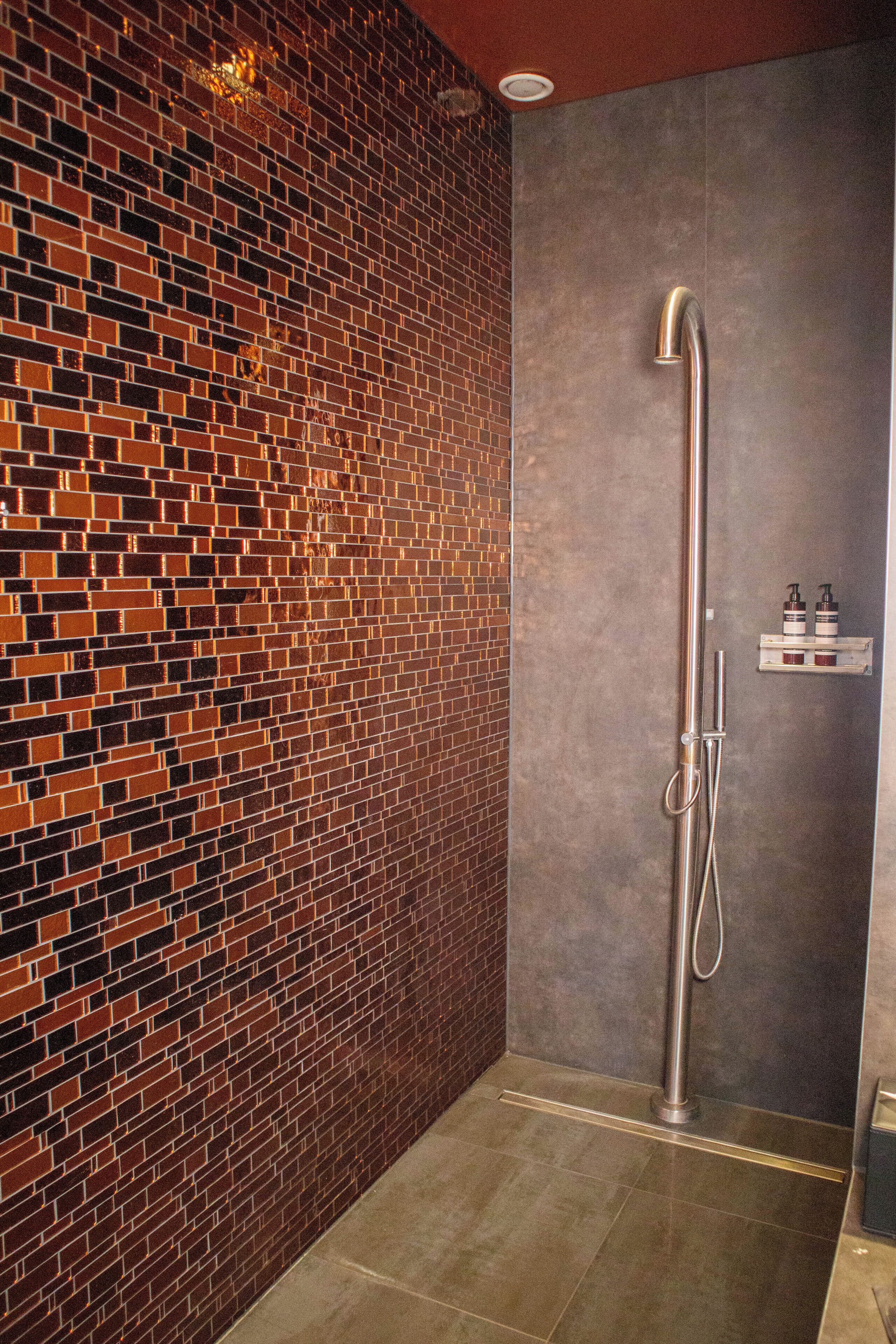 A modern bathroom with a brick-patterned wall, a shower with a handheld showerhead, and a small shelf with toiletries.