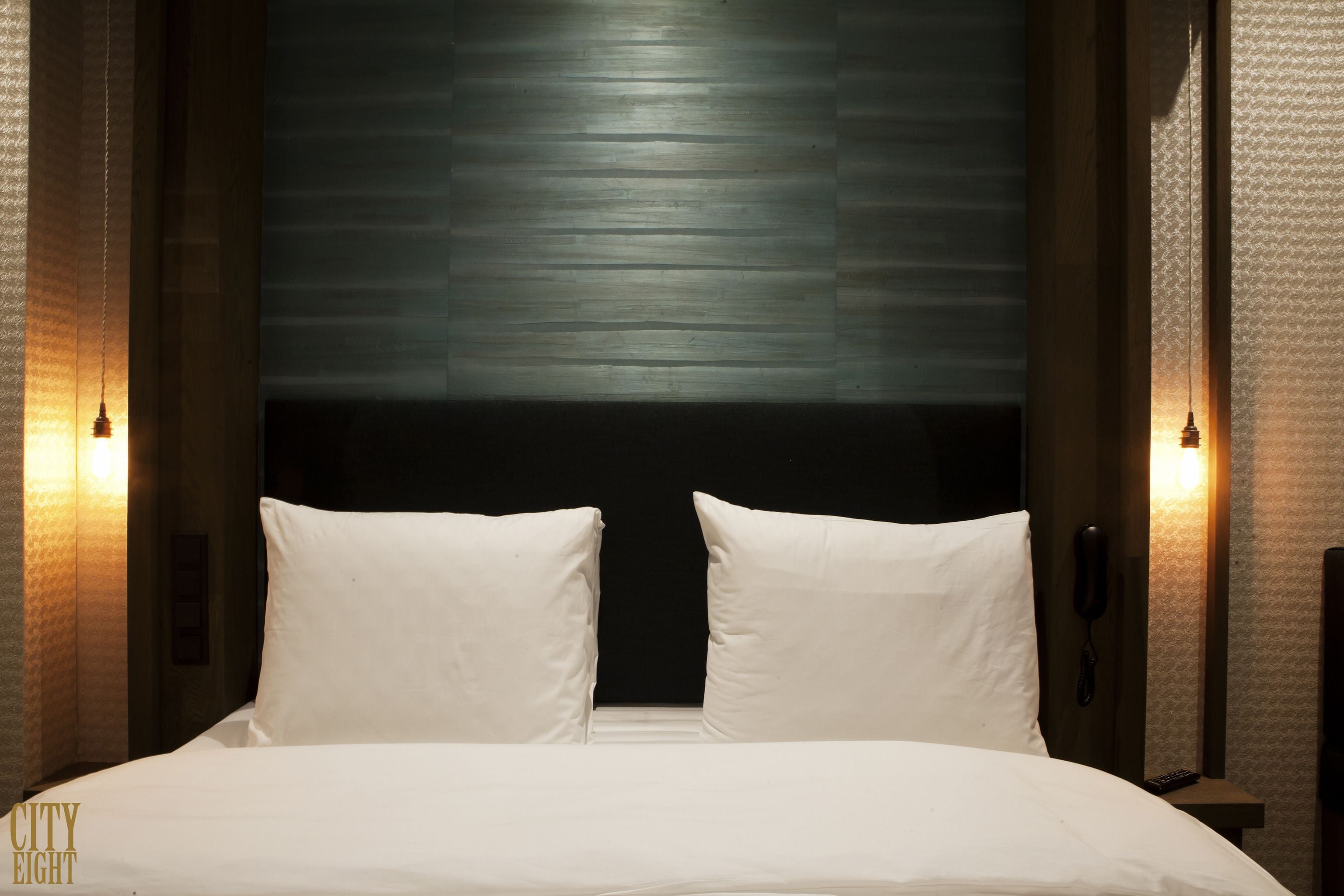 A hotel room with a large bed, two white pillows, a dark headboard, and two wall-mounted lamps.