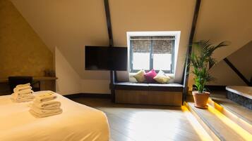 Una habitación de hotel con cama, un asiento junto a la ventana con cojines, un televisor de pantalla plana y una planta en maceta.