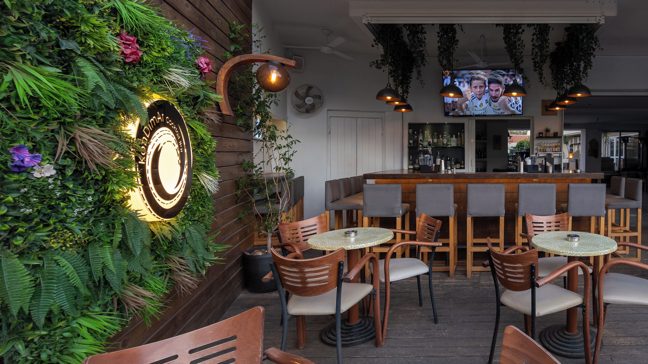 Un área de bar con muebles de madera, una pantalla de televisión y una pared decorada con plantas y un letrero.