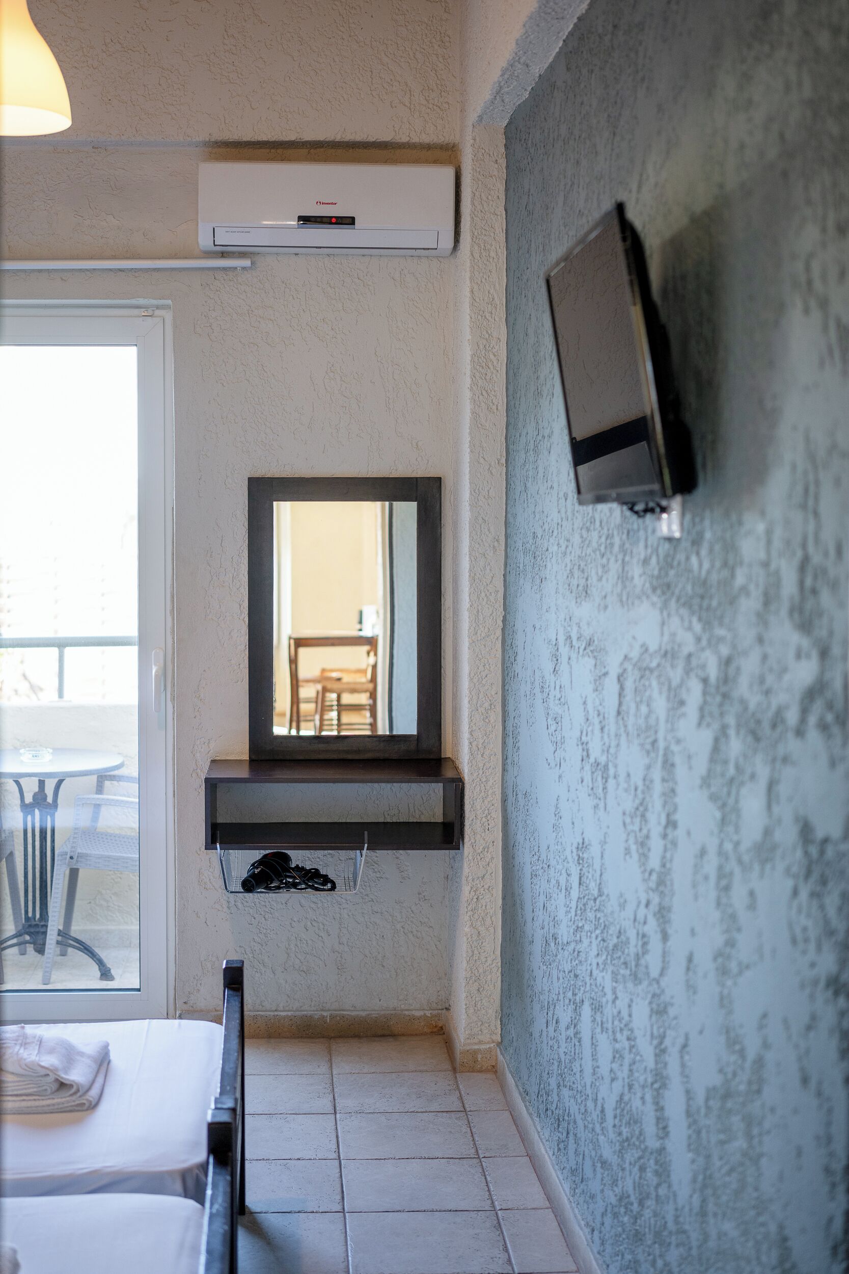 A hotel room with a bed, a bedside table, a mirror, a TV mounted on the wall, and an air conditioning unit above the mirror.