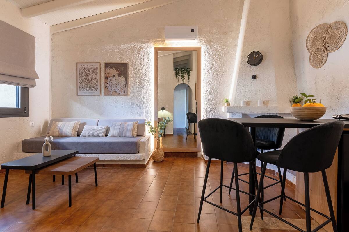 Un salon chaleureux avec un canapé, une table basse et un coin repas avec des chaises. On peut apercevoir la cuisine à travers une porte ouverte.