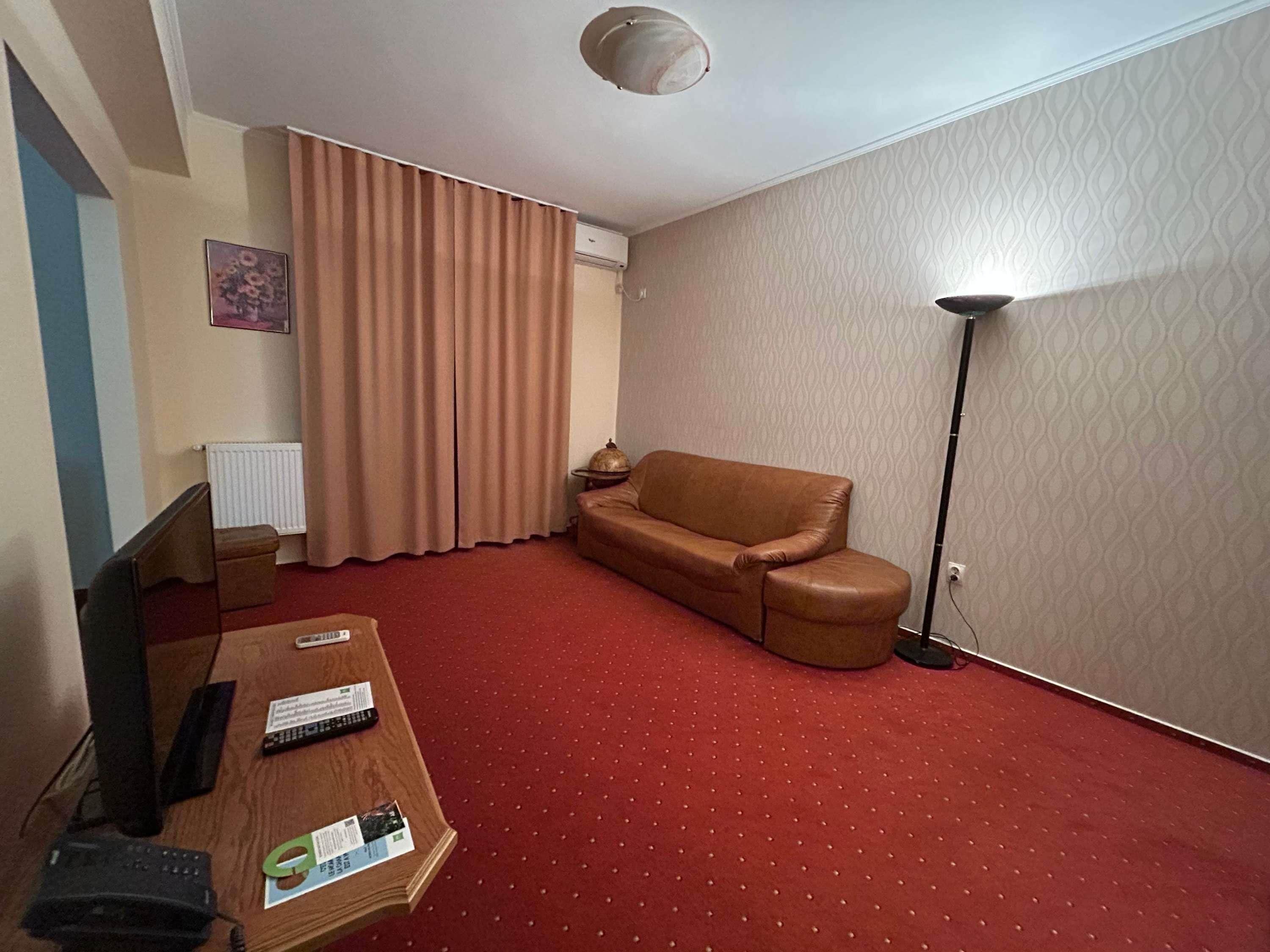 Une chambre d’hôtel avec une moquette rouge, un canapé en cuir marron, un bureau en bois avec des télécommandes, une lampe et un radiateur.