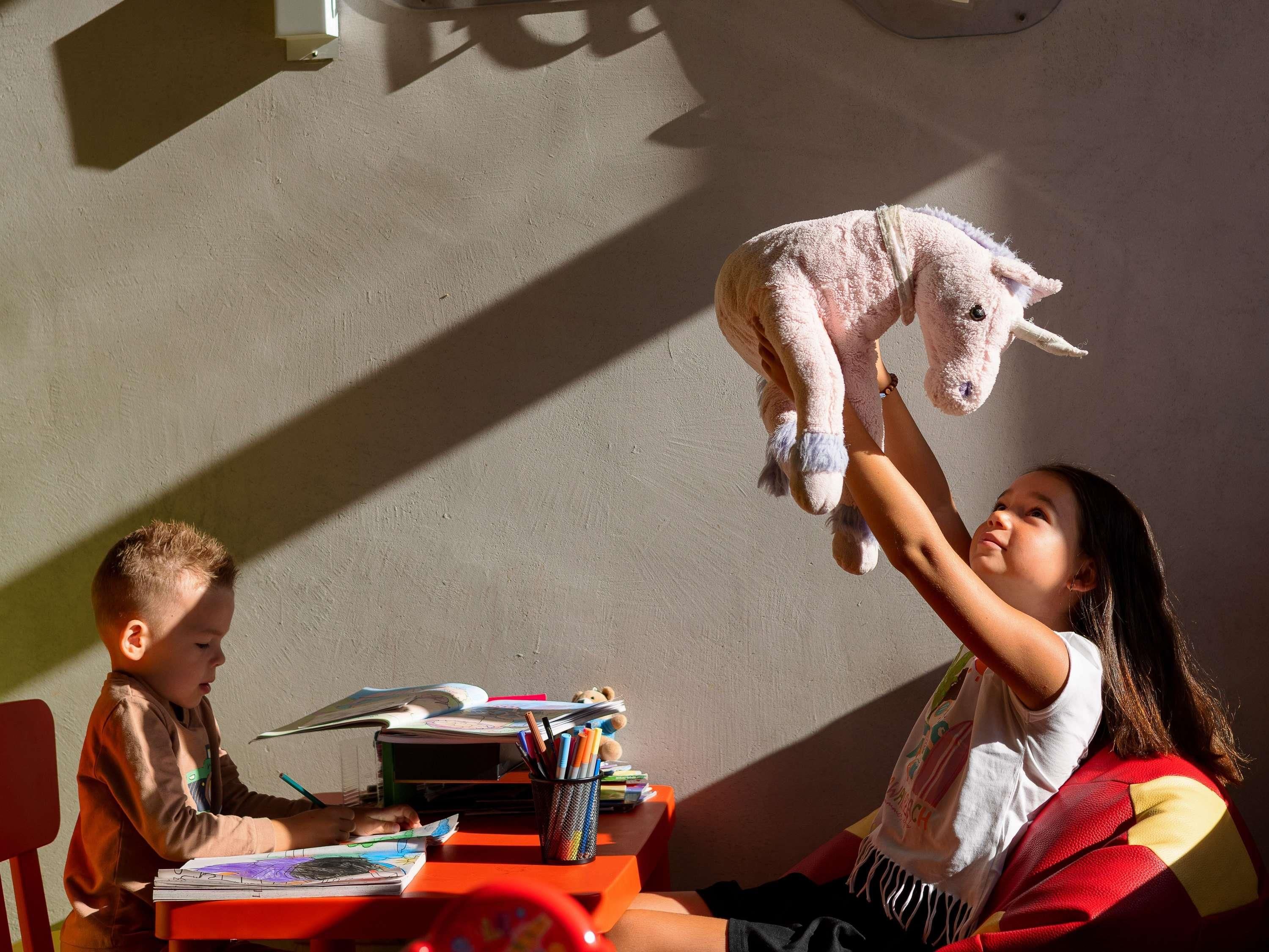 A child holding a stuffed toy above another child seated at a table with books and a cup of pens.