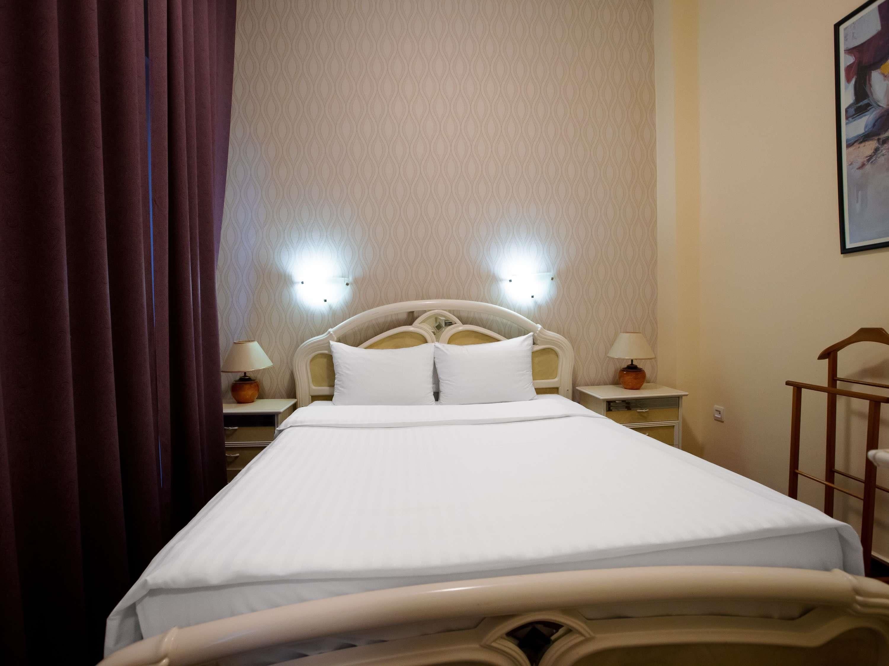 A hotel room with a bed, two bedside tables with lamps, a wall-mounted light fixture, and a curtain.