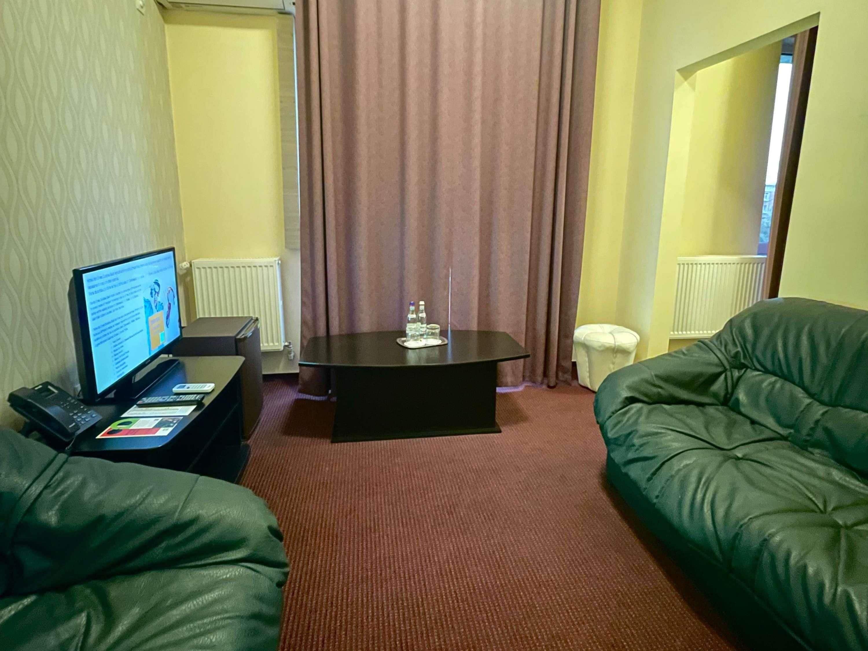 A hotel room with a desk, a computer, a green sofa, a small table with a glass, and a window with curtains.