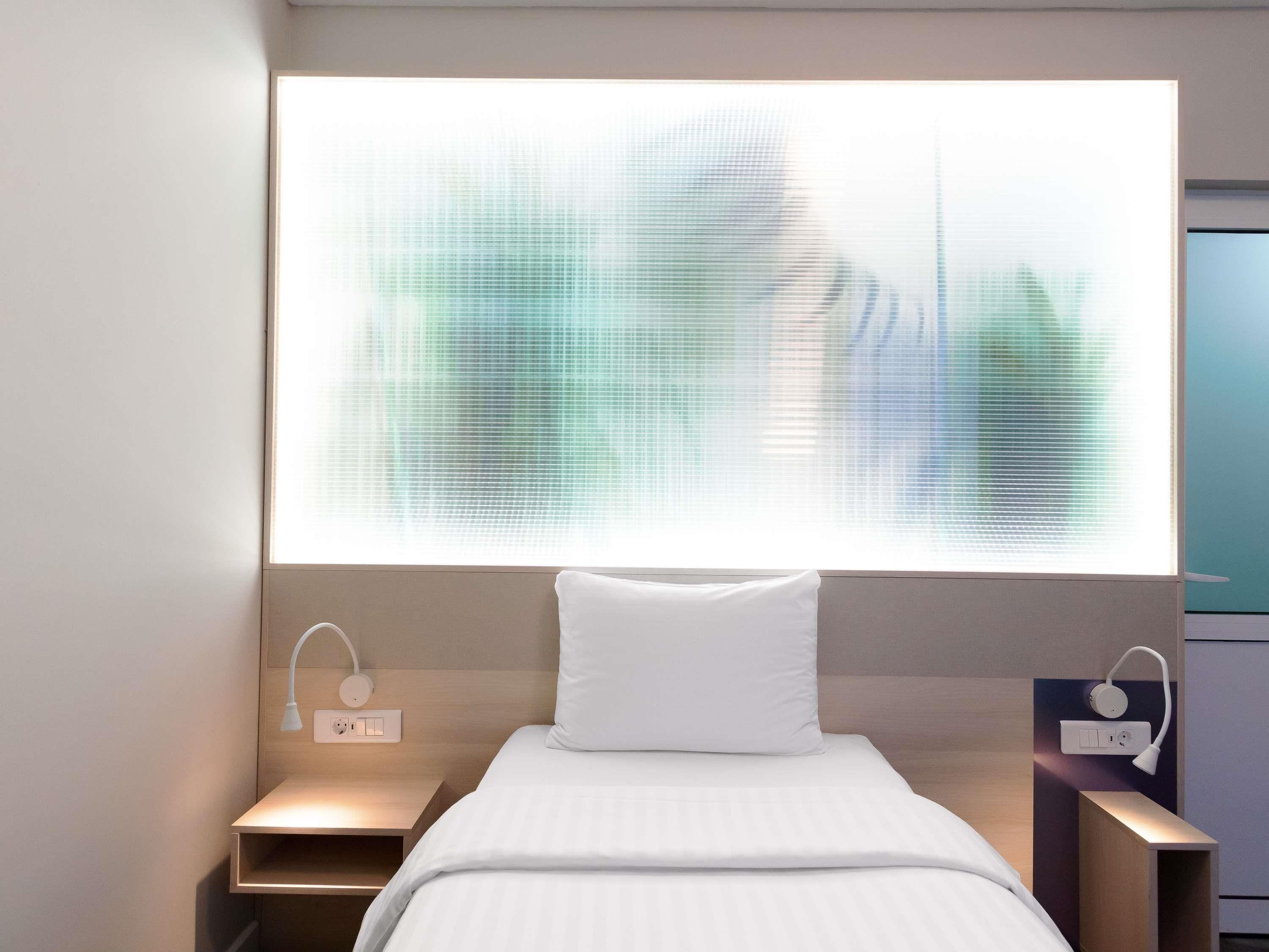 A modern hotel room with a large window, a bed with white bedding, bedside tables, and a wall-mounted light fixture.