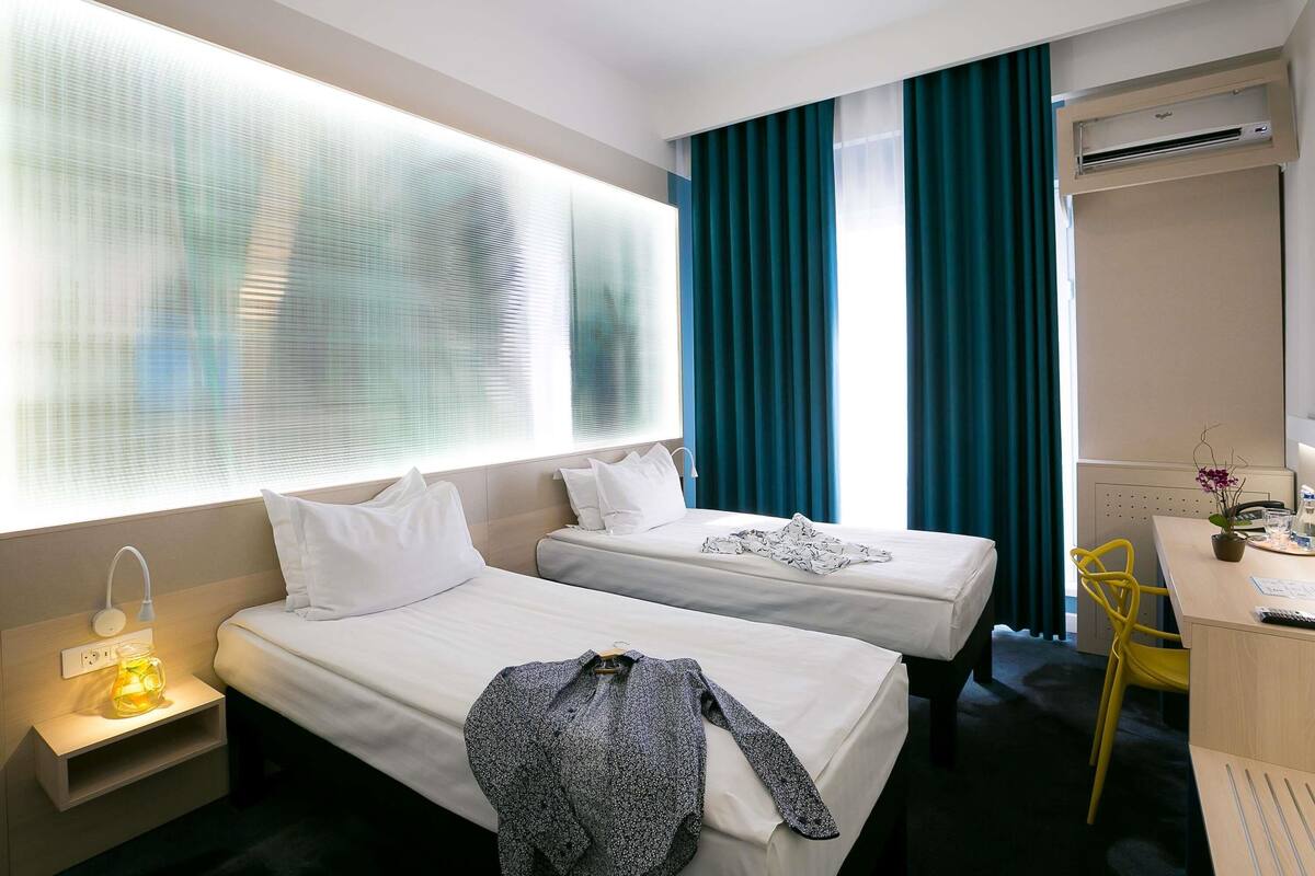 Ein Hotelzimmer mit zwei Betten, einem Schreibtisch mit Stuhl, einer Klimaanlage und einem Fenster mit Vorhängen.