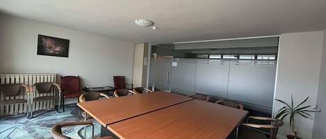 Un salón de conferencias con una larga mesa y sillas, un radiador, un cuadro y una planta en maceta.