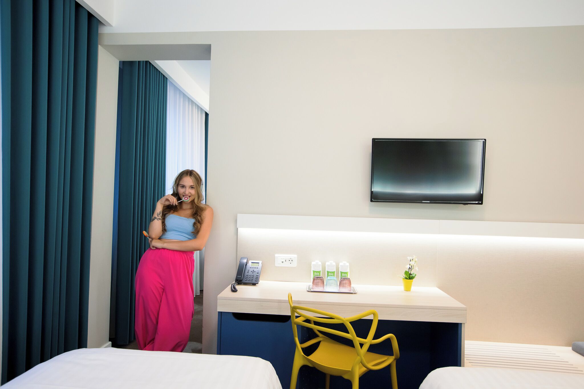 A hotel room with a bed, a desk with a chair, a TV mounted on the wall, and a window with curtains.