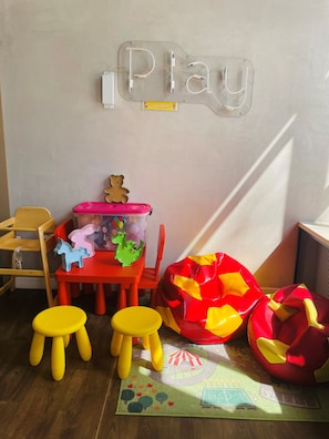 Een speelruimte voor kinderen met een bord waarop 'Play' staat, een rode tafel, gele krukjes, een opbergmeubel voor speelgoed in roze en groen, en een rode loungestoel.