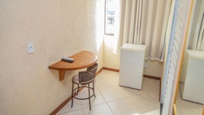 A small room with a wooden table, a wicker chair, a white refrigerator, and a window with curtains. - Pousada Ondas do Forte (Cabo Frio)