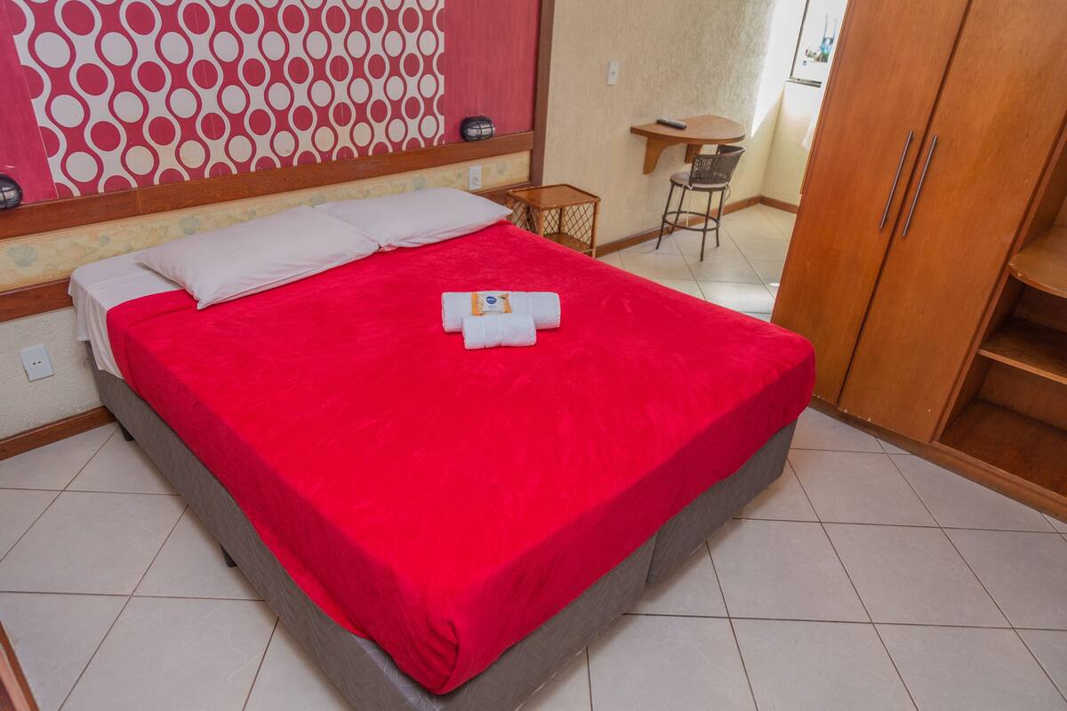 A hotel room with a large bed, a wooden wardrobe, a small table, and a chair.