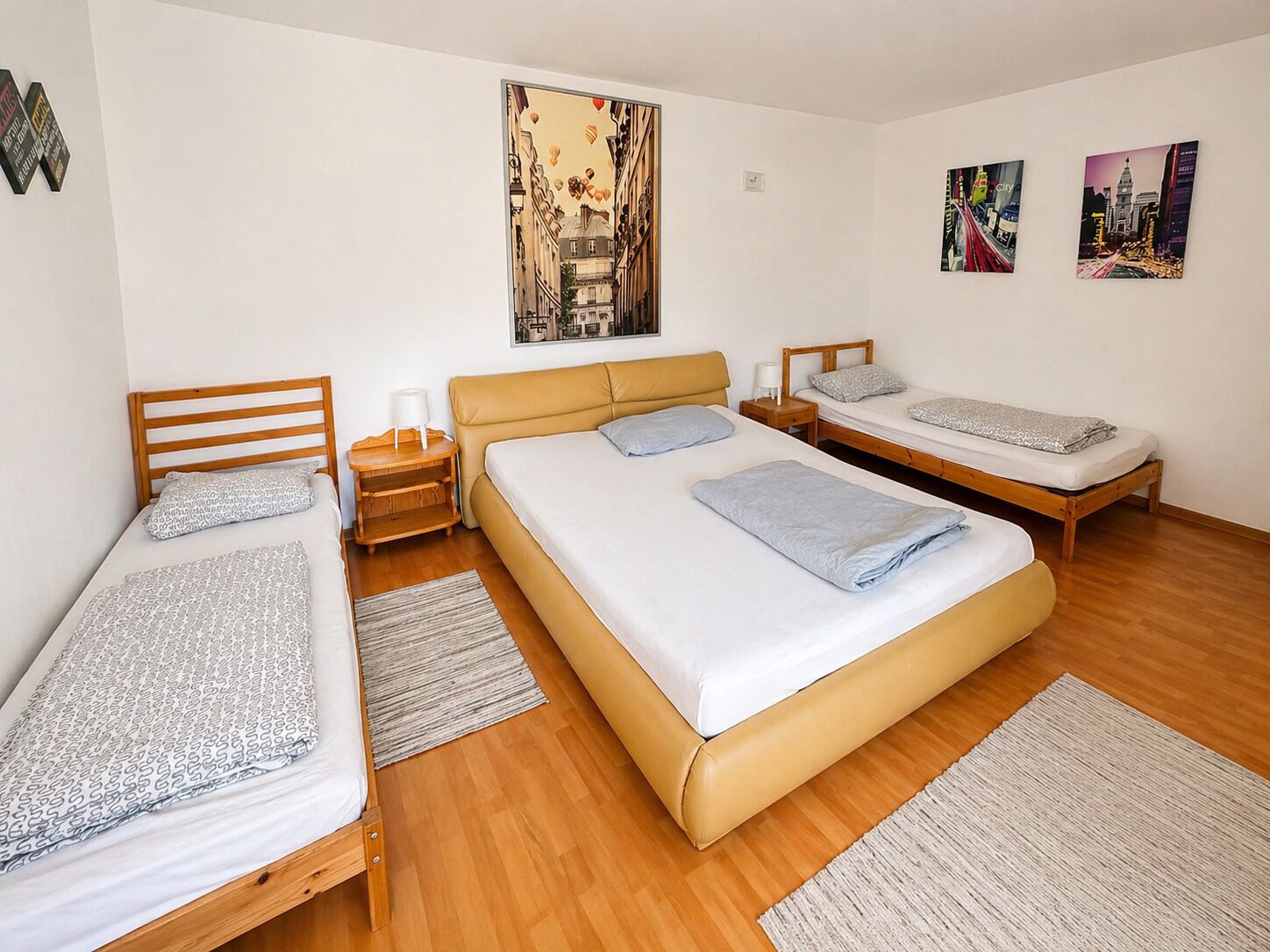 A room with three beds, a wooden floor, and a cityscape painting on the wall.