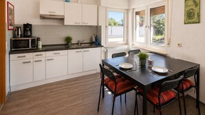 A modern kitchen with white cabinets, a dining table with chairs, and a window providing a view of greenery outside. - Apartment Hope Inn (Gross-Zimmern)