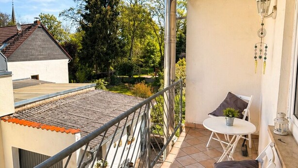 A balcony with a metal railing, a small table with a chair, a potted plant, and a decorative lantern. - Apartment Hope Inn (Gross-Zimmern)