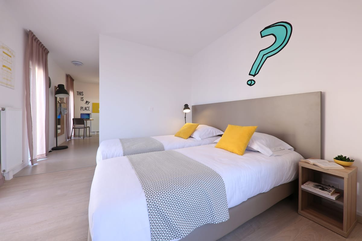 A modern hotel room with two beds, a bedside table with a lamp, and a small wooden side table with a plant and books.
