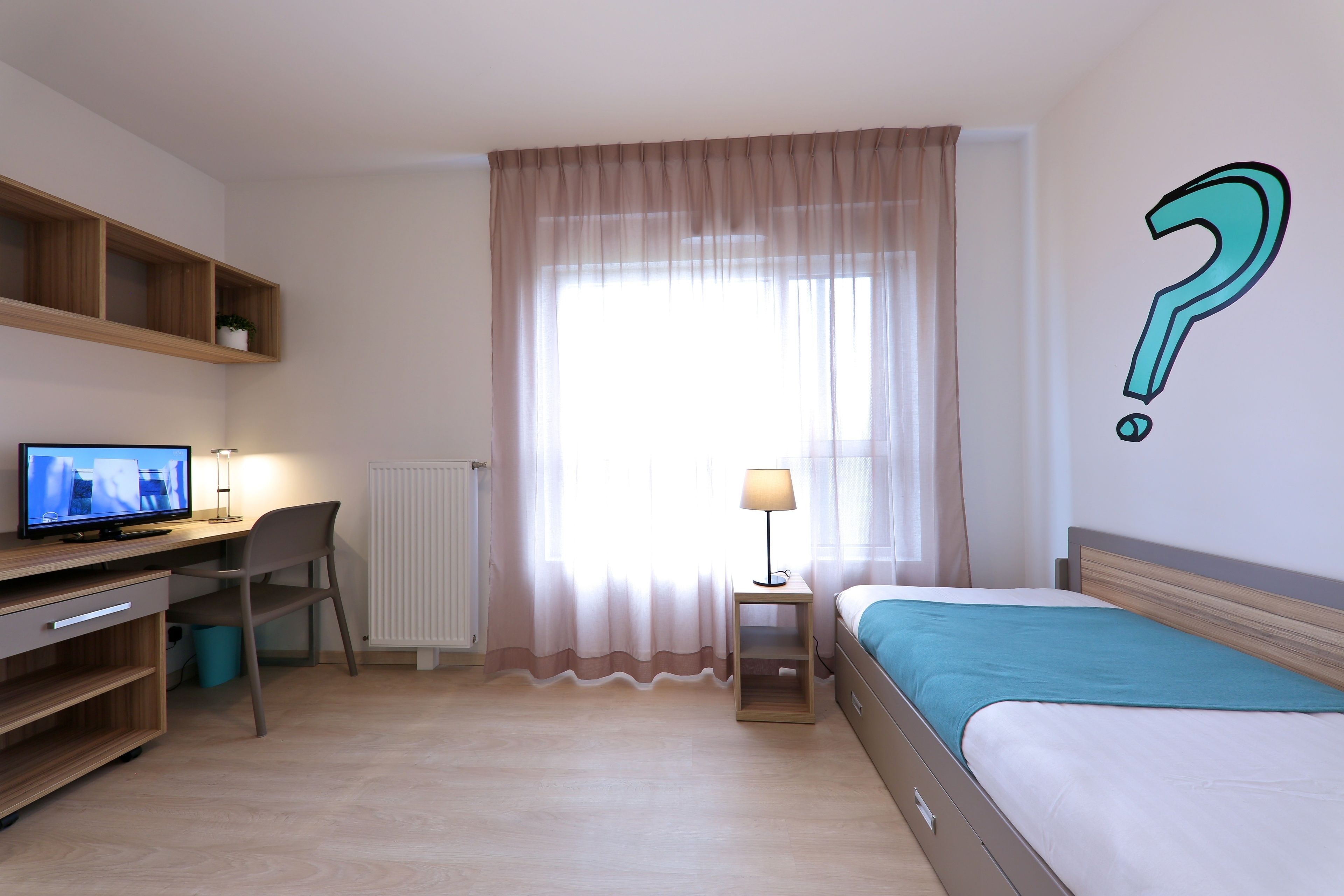 A hotel room with a bed, a desk with a computer, a chair, a lamp, and a window with sheer curtains.