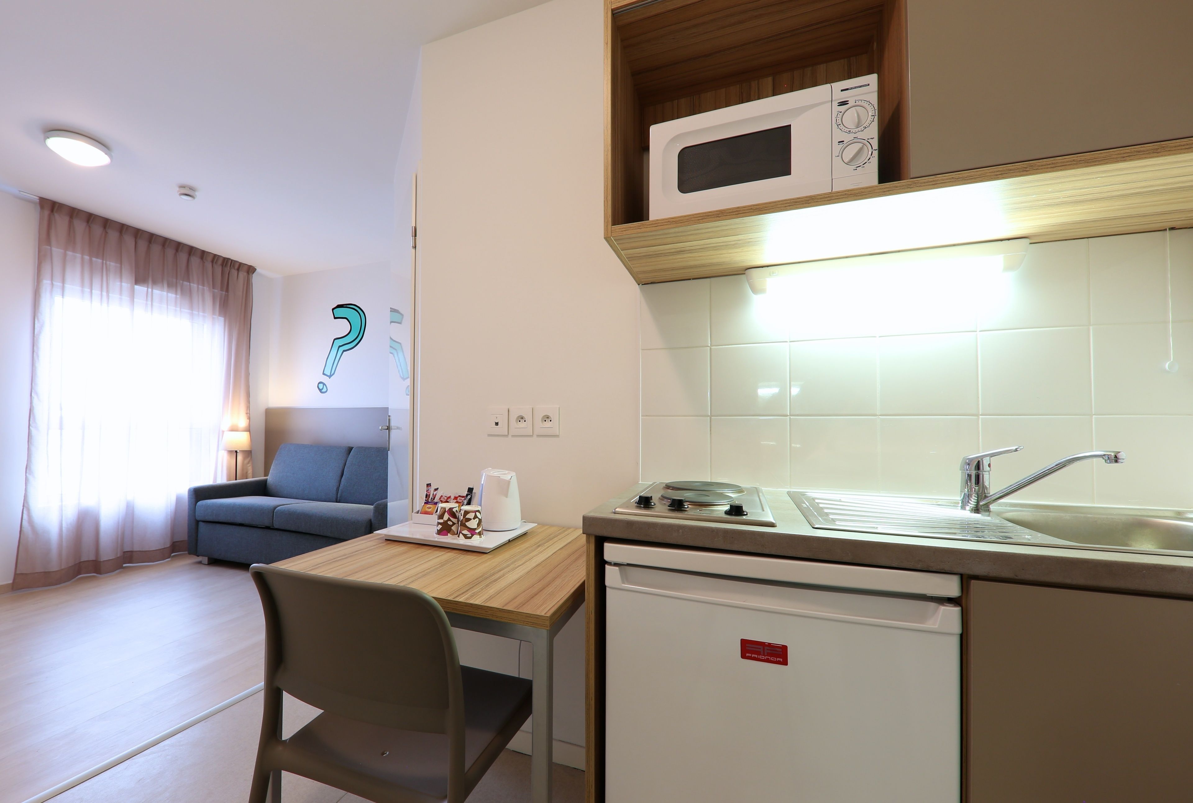 A compact kitchen with a microwave, sink, and a small refrigerator. There is a dining area with a table and a chair.