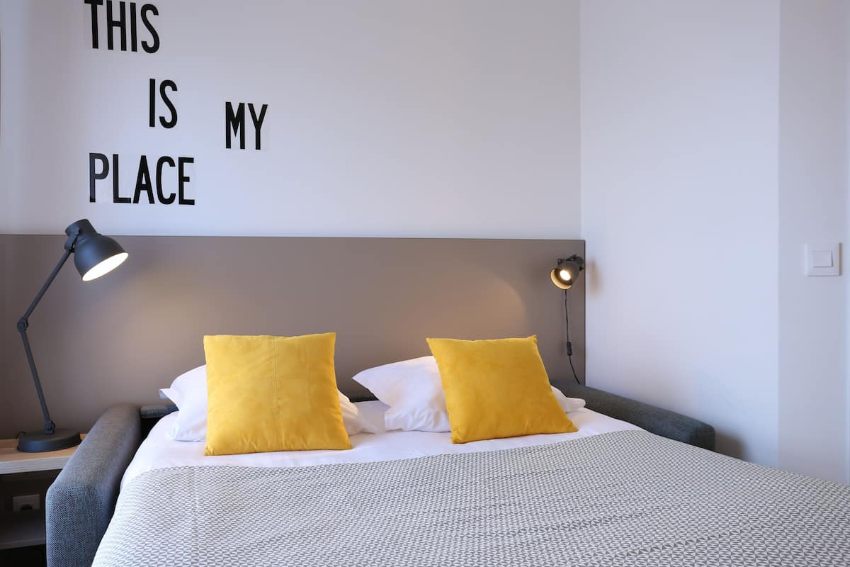 A neatly made bed with two yellow pillows, a grey headboard, and a wall sign that reads "THIS IS MY PLACE".
