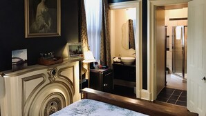 A hotel room with a bed, a painting, a lamp, a bathroom with a sink and mirror, and a shower.