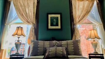 Un dormitorio con una cama, dos lámparas de noche, un retrato enmarcado en la pared y cortinas.