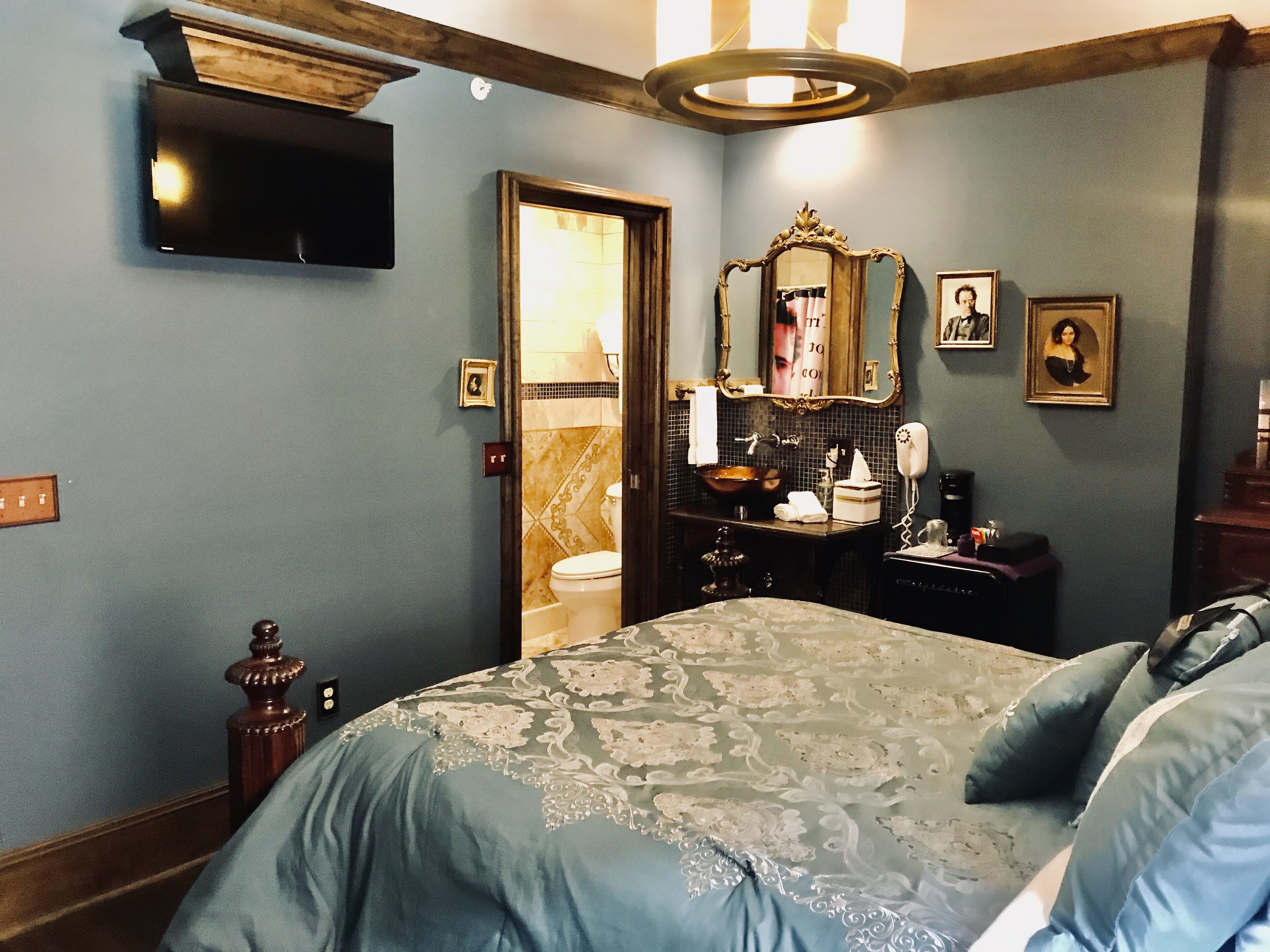 A bedroom with a large bed, a wooden headboard, a television mounted on the wall, a mirror with a decorative frame, and a small bedside table with a lamp.