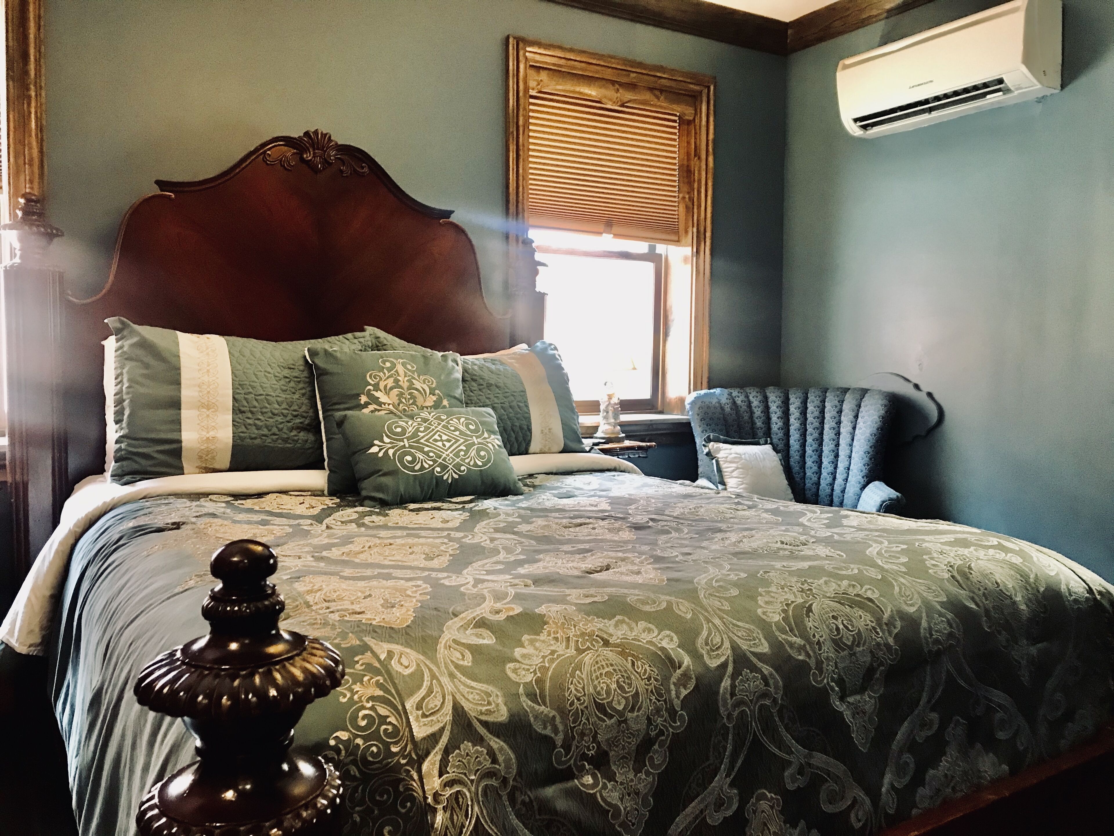 Une chambre avec un grand lit, une tête de lit en bois, une fenêtre avec des stores, une chaise et un appareil de climatisation.