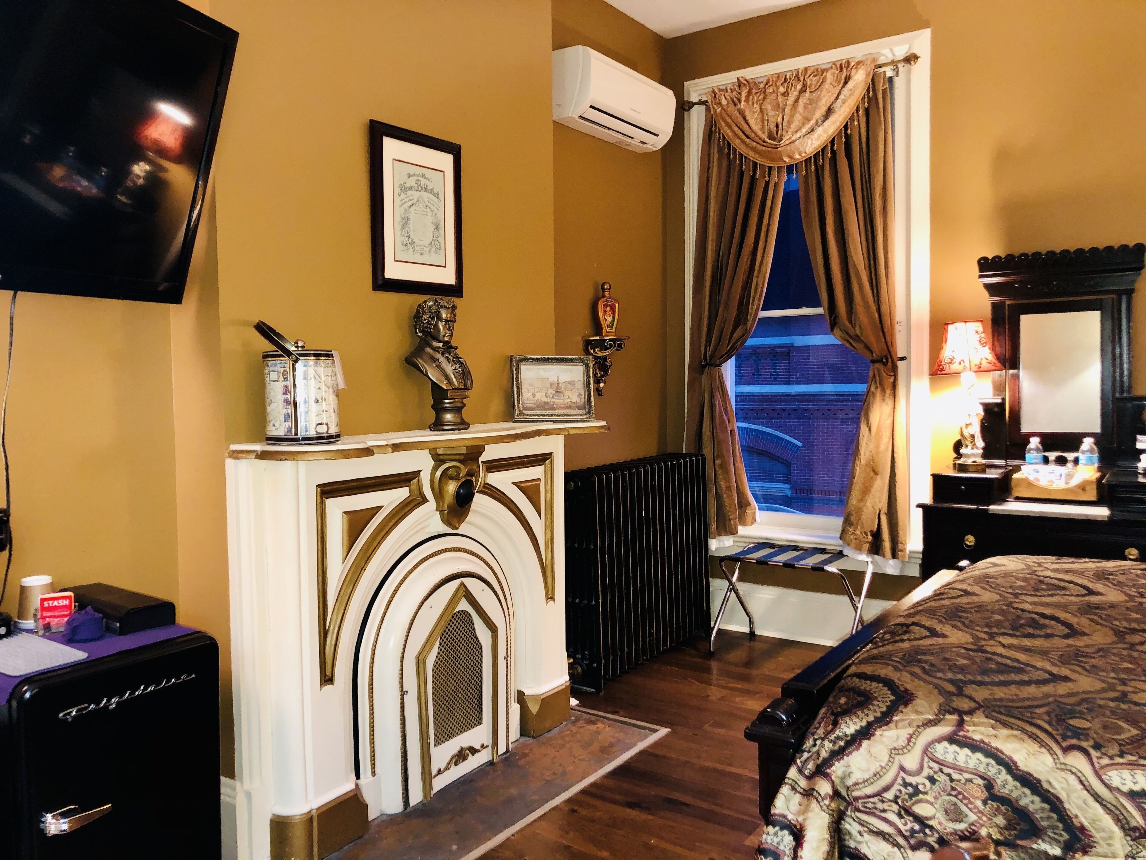 A hotel room with a fireplace, a television, a radiator, a window with curtains, and a bed with a patterned bedspread.