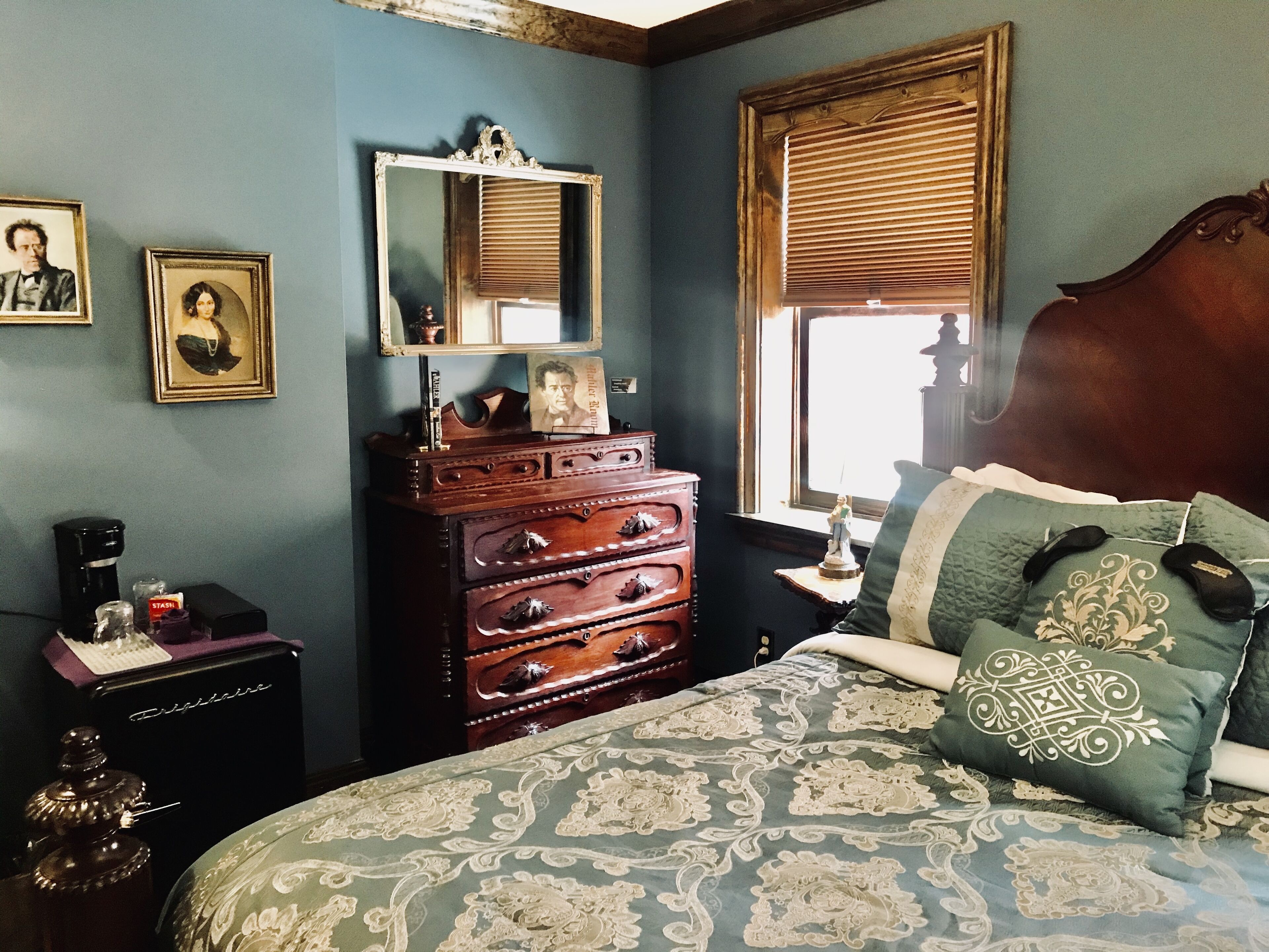 Un dormitorio con un aparador de madera, una cama con ropa de cama estampada, una mesita de noche con lámpara y una ventana con persianas.