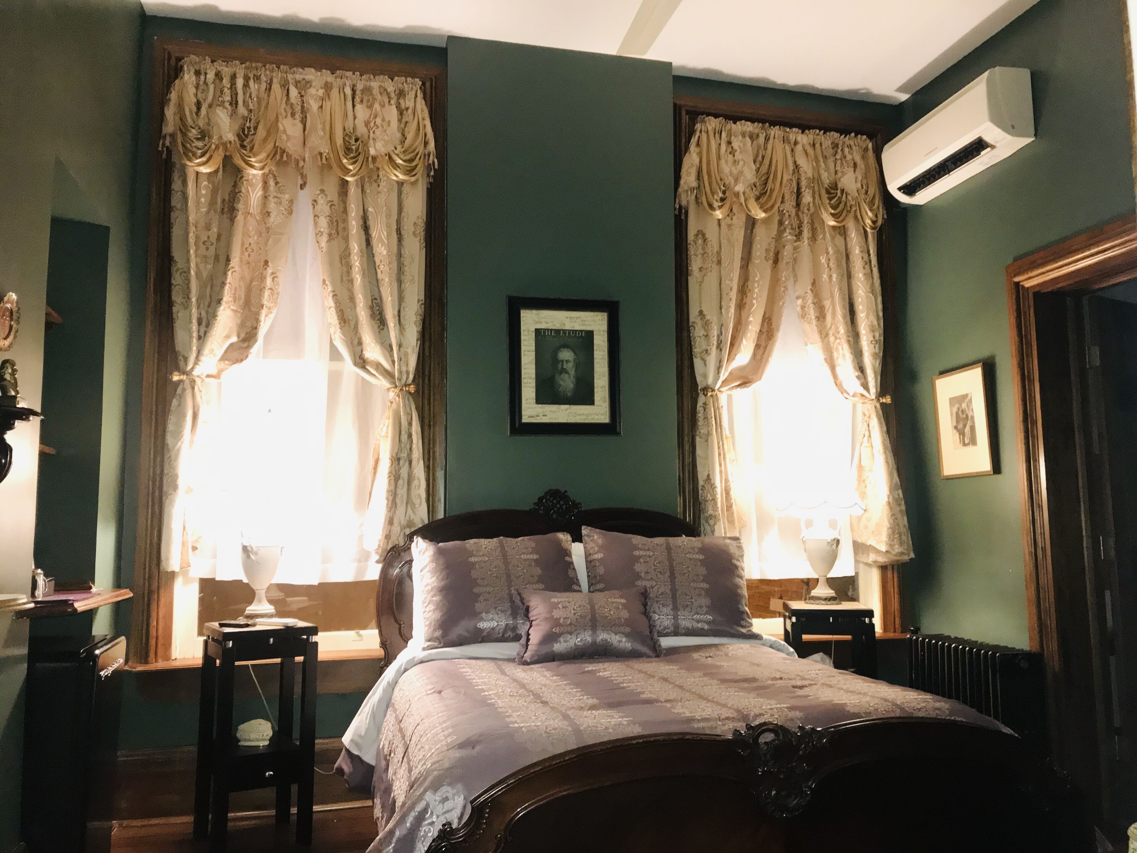 Ein Schlafzimmer mit einem großen Bett, zwei Nachttischen, einem Schminktisch und einem gerahmten Bild an der Wand. An der Wand ist außerdem eine Klimaanlage angebracht.