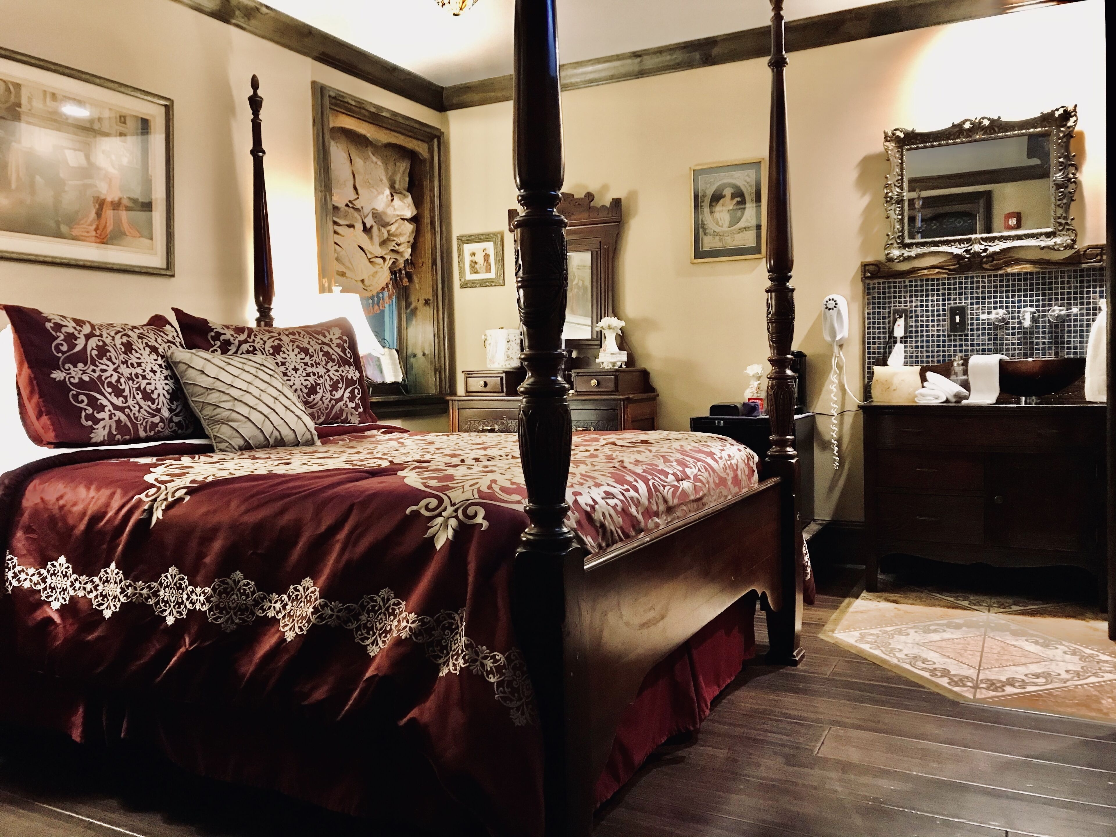 Una cama con dosel con edredón rojo y blanco, una mesita de noche de madera con lámpara, un aparador con espejo y un baño con lavamanos y espejo.