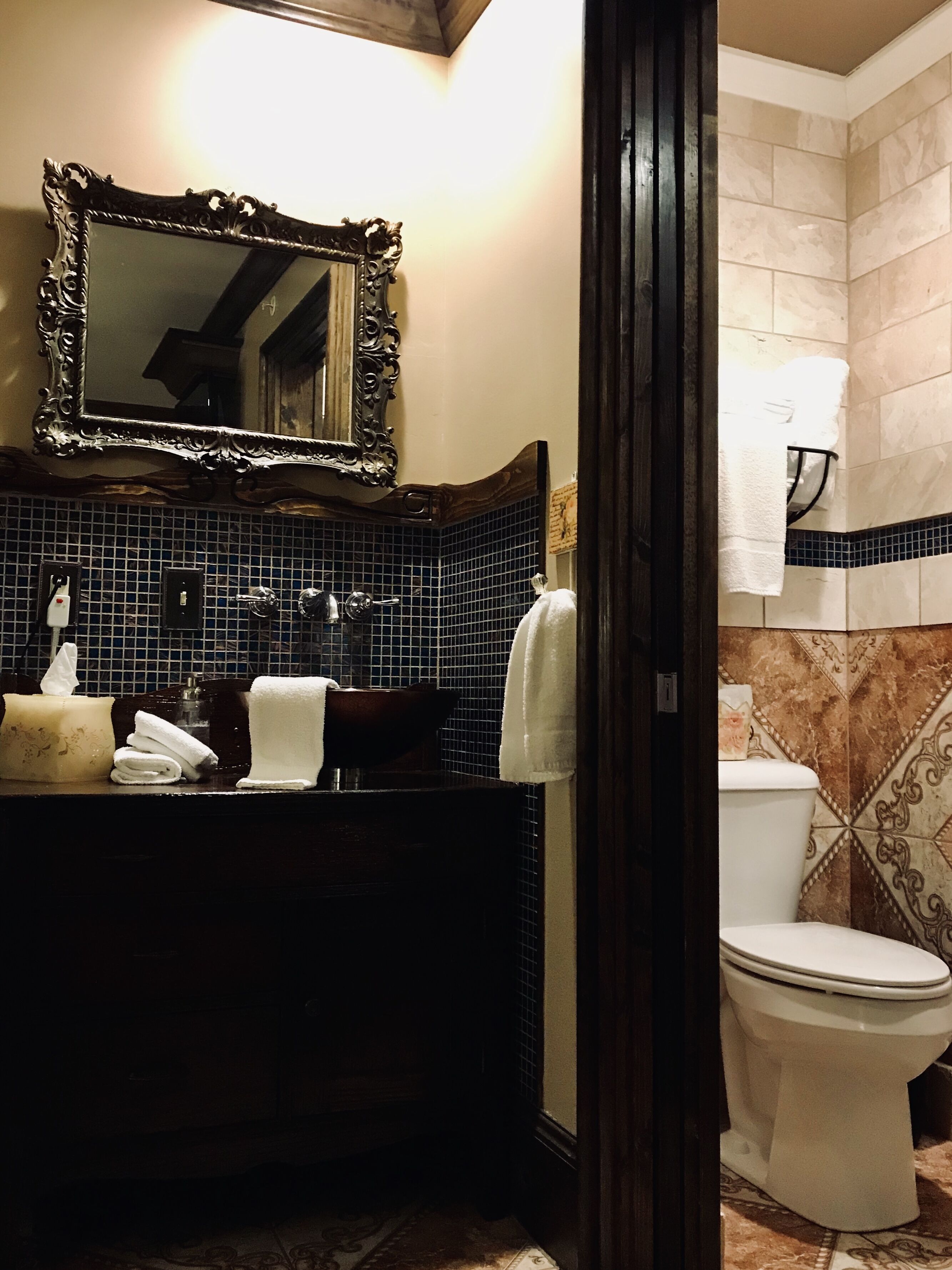 Un baño con un mueble de tocador de madera oscura, un espejo con marco decorativo, un inodoro blanco y un salpicadero de azulejos.